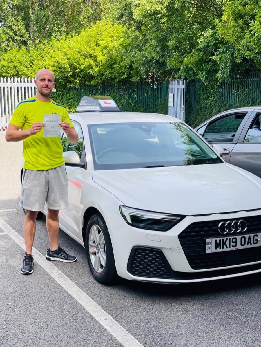 Holding up a pass certificate in a lime sports top and grey shorts, a learner stands beside the tuition car in bright sunshine.