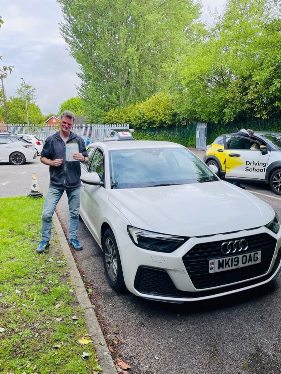 Grinning on a grassy verge in a zip-up hoodie and jeans, a learner shows a pass certificate, another driving-school car behind.