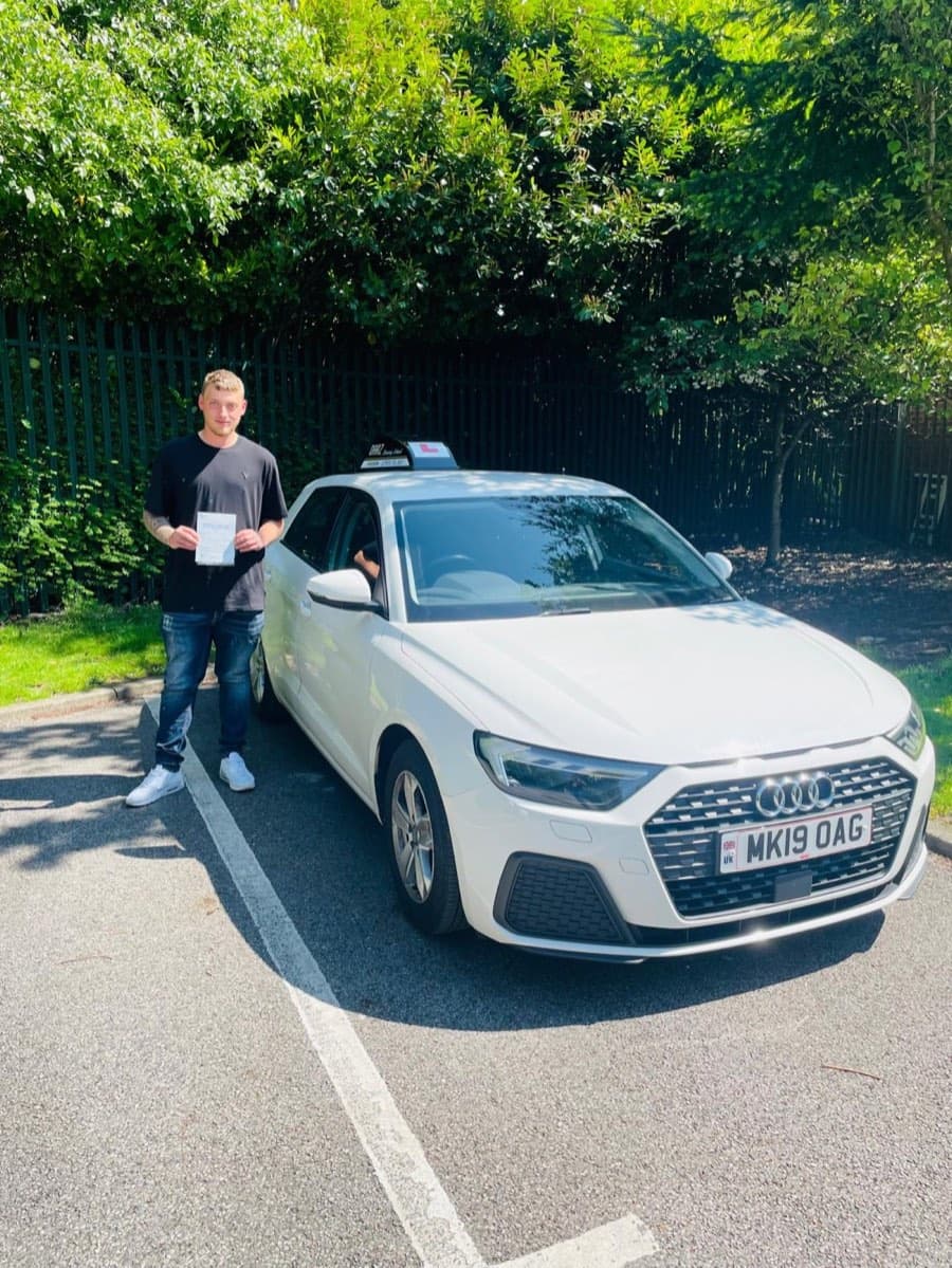In a black t-shirt, jeans and white trainers, a learner holds a pass certificate by the car in summer sunshine.
