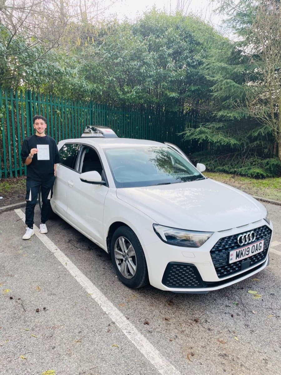 Beside the white Audi on a grey winter day, a learner in black sportswear raises a pass certificate towards the camera.