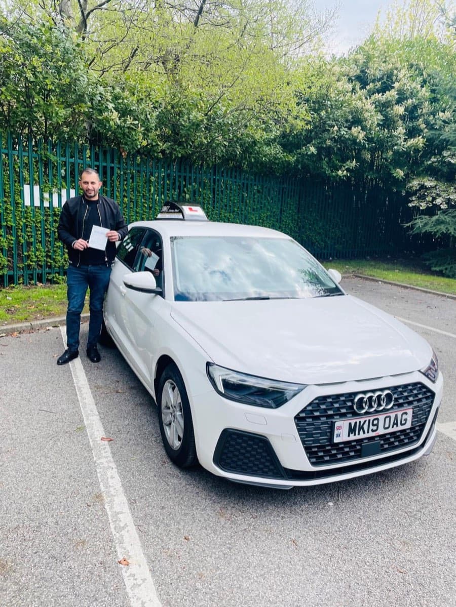 Smiling in a black bomber jacket and jeans, a learner holds a pass certificate by the car against green railings.