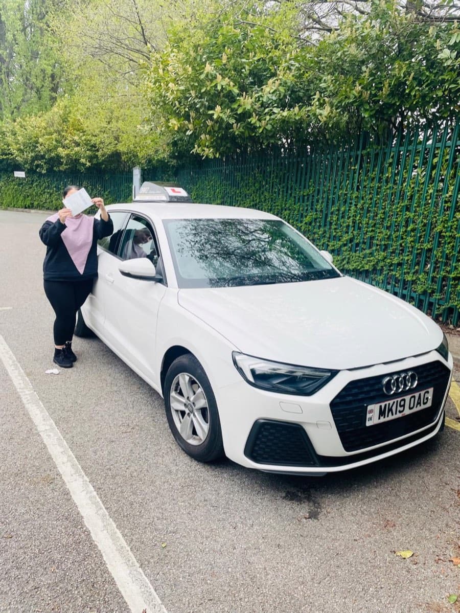 A learner in a black and lilac hoodie holds a pass certificate up in front of their face beside the car.
