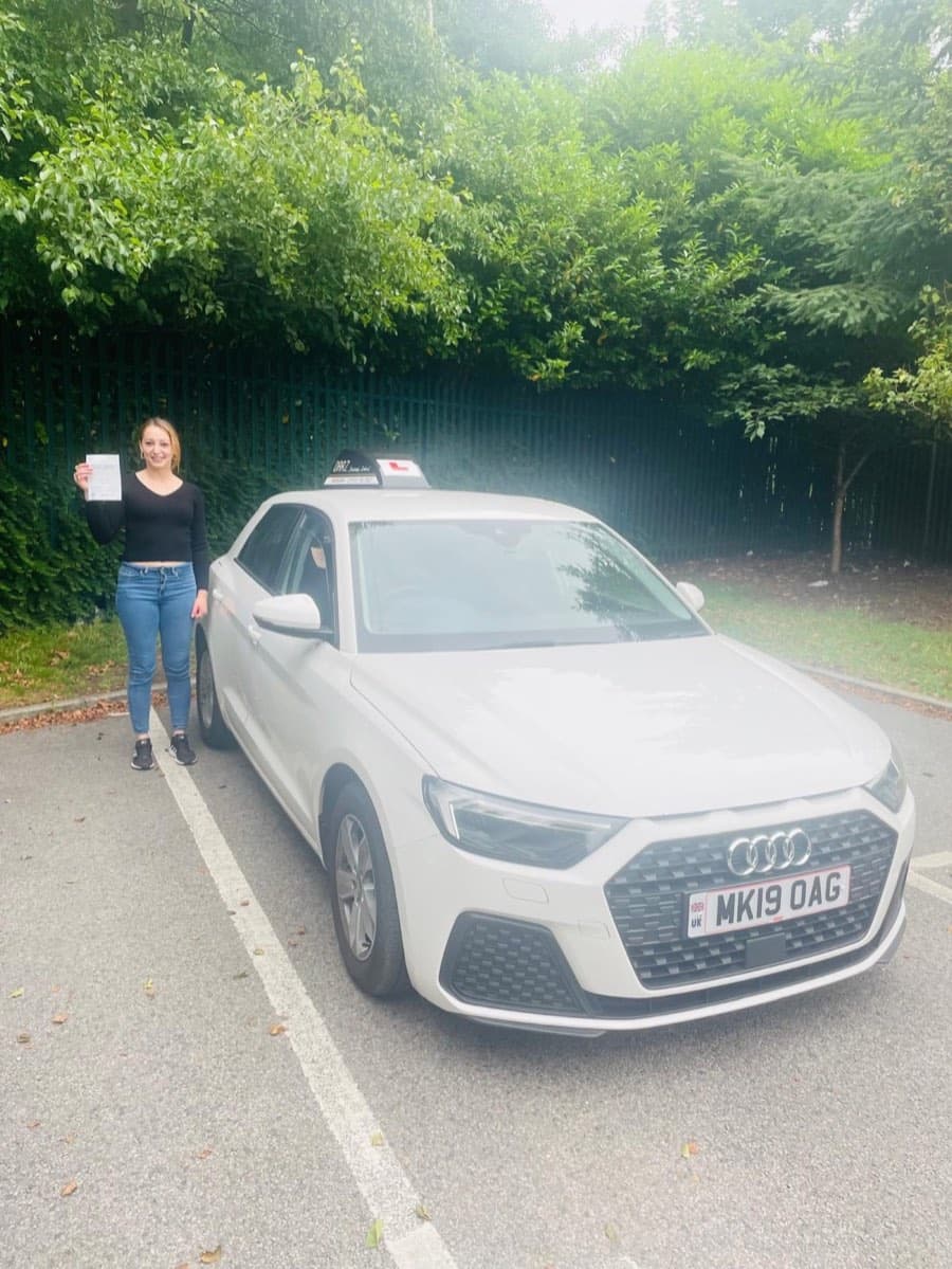 Arm raised high with the certificate on an overcast day, a learner in a black top and jeans stands by the bonnet.