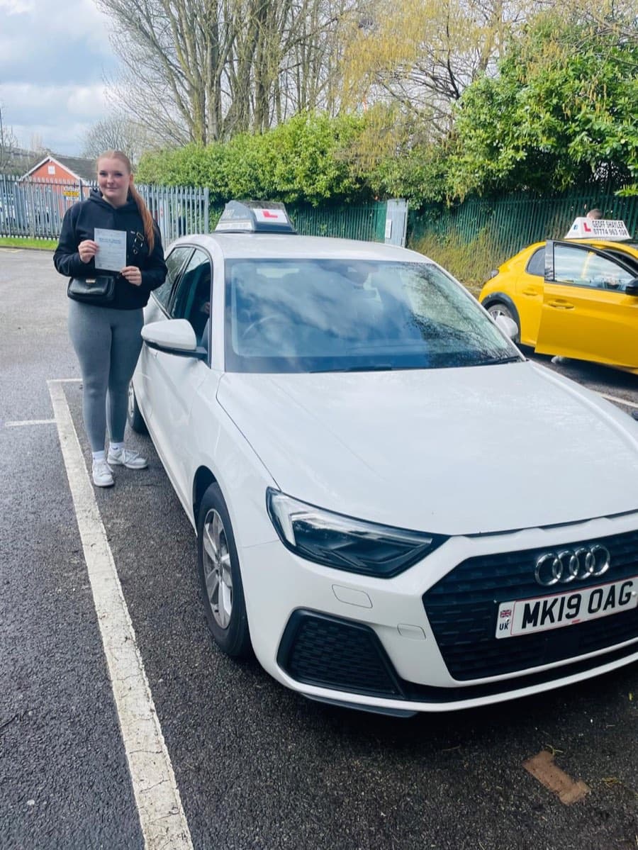 A learner in black hoodie, grey leggings and white trainers holds the certificate, another tuition car parked behind.