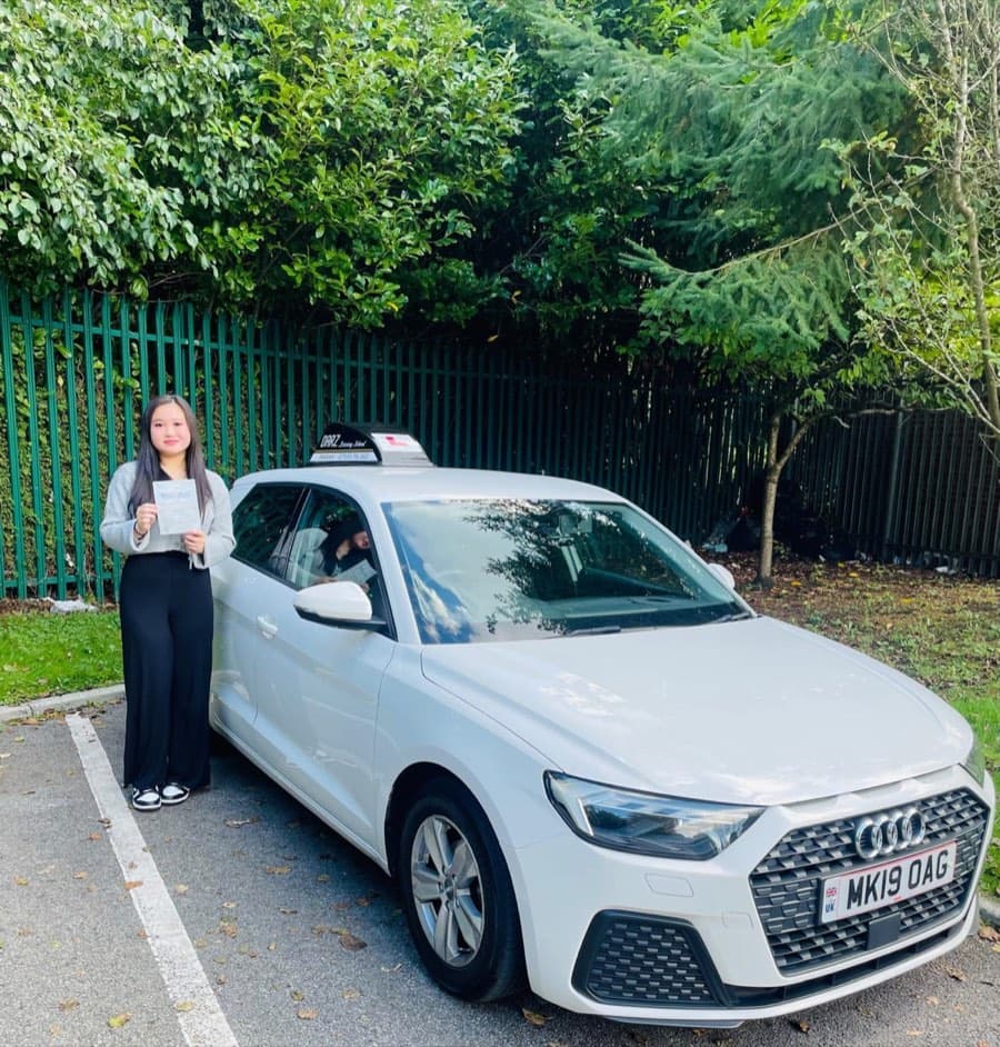 Holding the certificate in both hands beside the tuition car in autumn, a learner wears a grey jumper and wide-leg trousers.