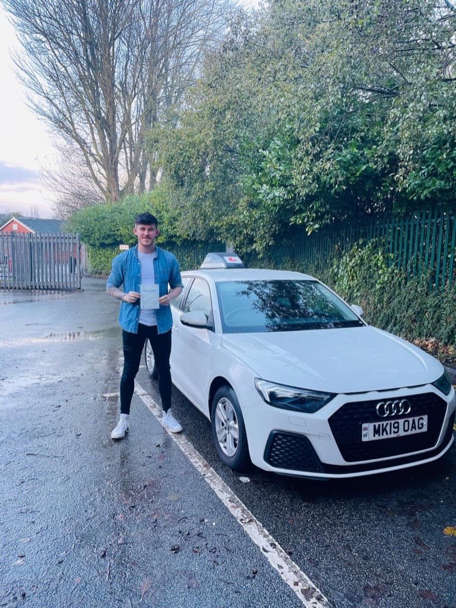 On a rain-soaked road beside the white tuition car, a learner in an open denim shirt and white trainers holds a certificate.