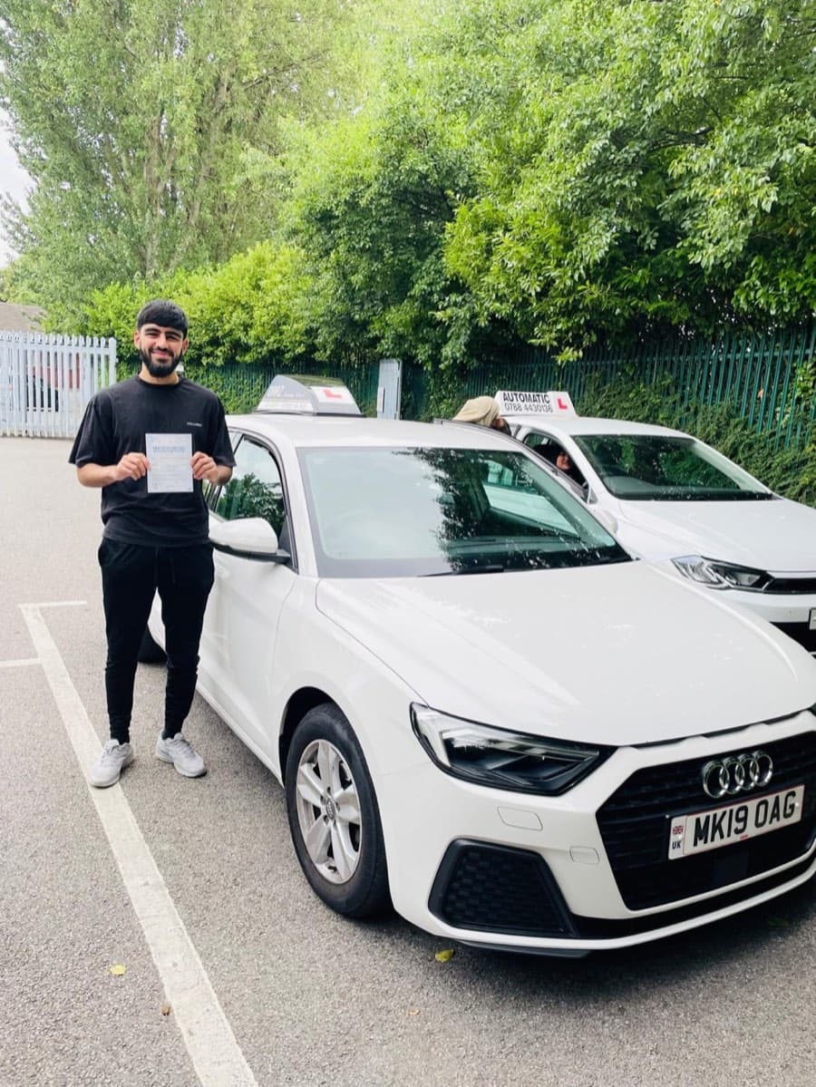 A learner dressed head to toe in black holds a certificate, with a second tuition car parked alongside in sunshine.