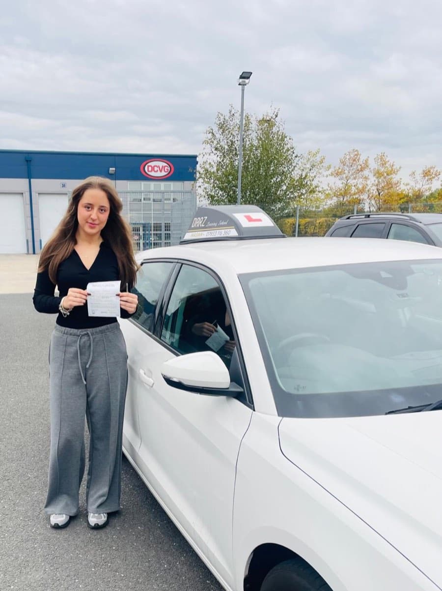 A learner in a black top and grey joggers holds a certificate beside the white tuition car outside an industrial unit.