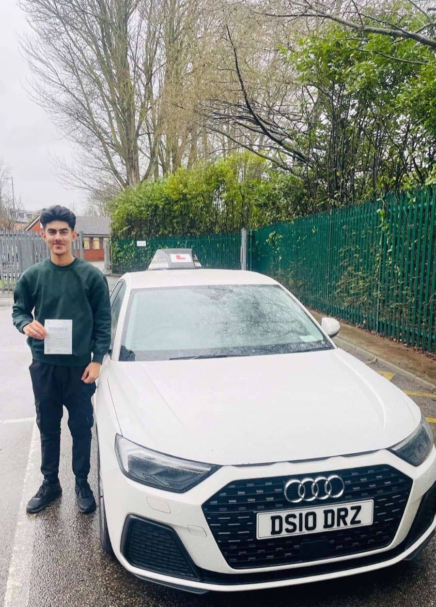 Dressed in a dark green sweatshirt and black trousers, a learner displays a certificate beside the tuition car on damp tarmac.