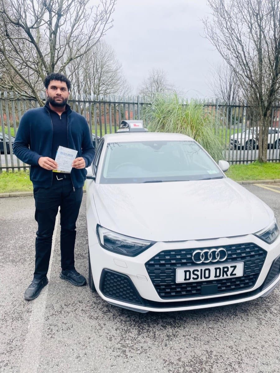 Wrapped in a navy zip-up jacket, a learner holds a certificate by the tuition car amid bare winter trees.
