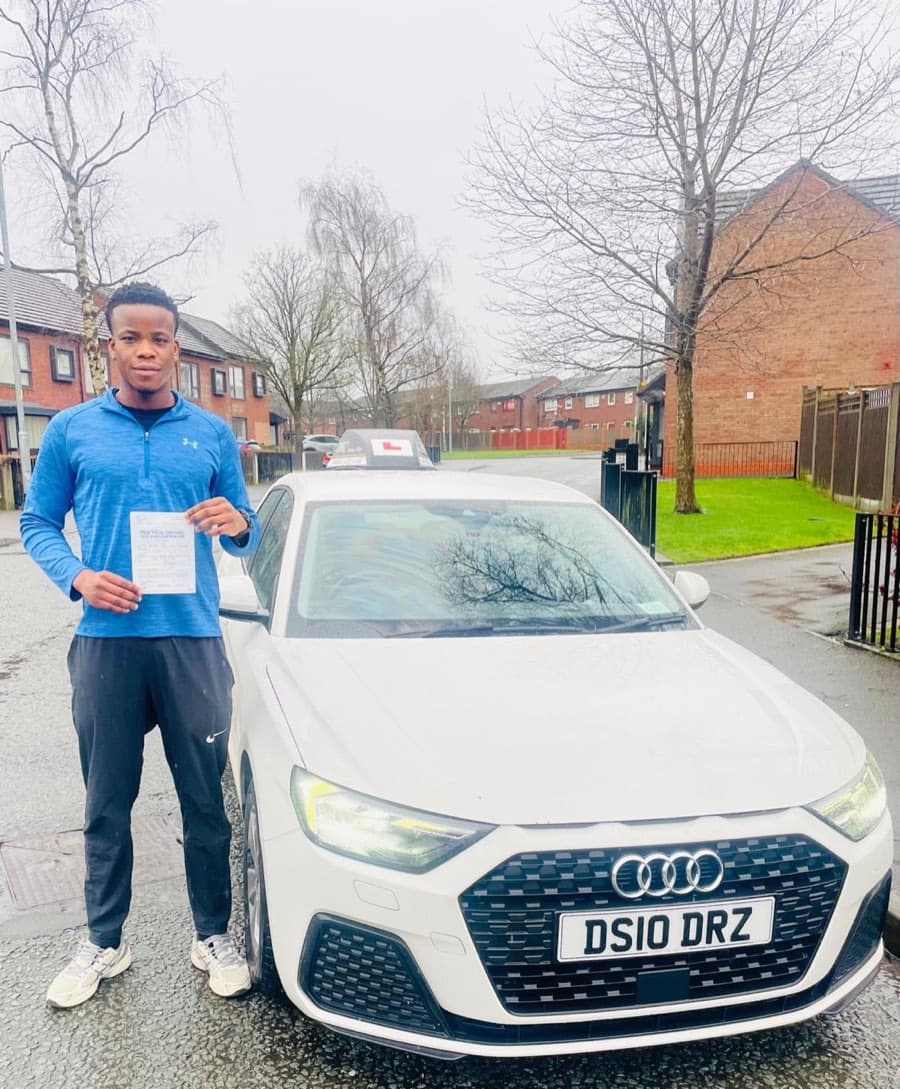 Holding a certificate beside the tuition car, a learner in a blue sports top stands on a wet residential street.
