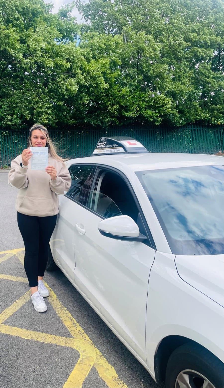 On a breezy day, a learner in a beige hoodie and black leggings holds a certificate beside the white tuition car.