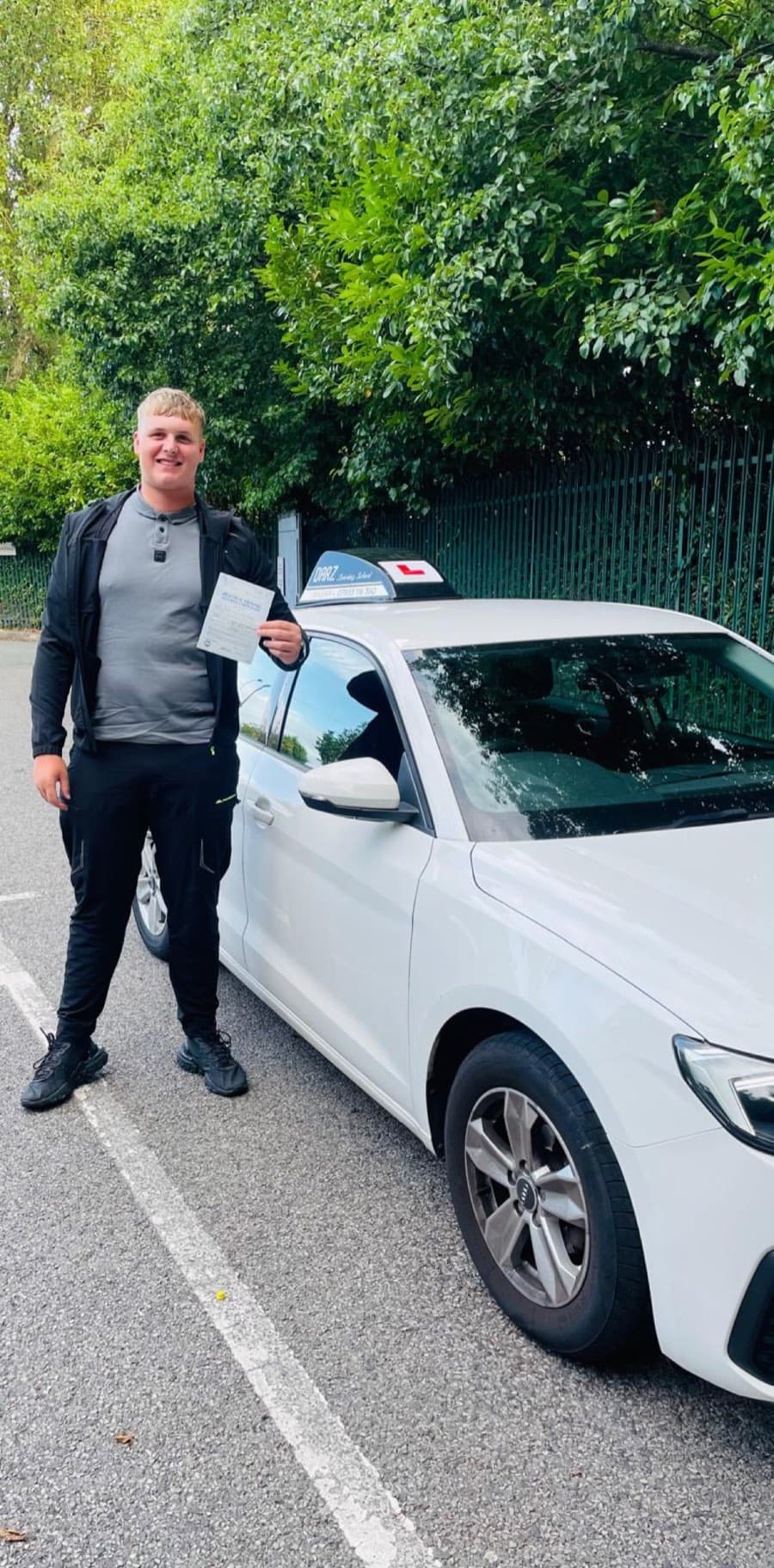 Wearing a black jacket over a grey polo shirt, a learner stands by the tuition car holding a pass certificate.