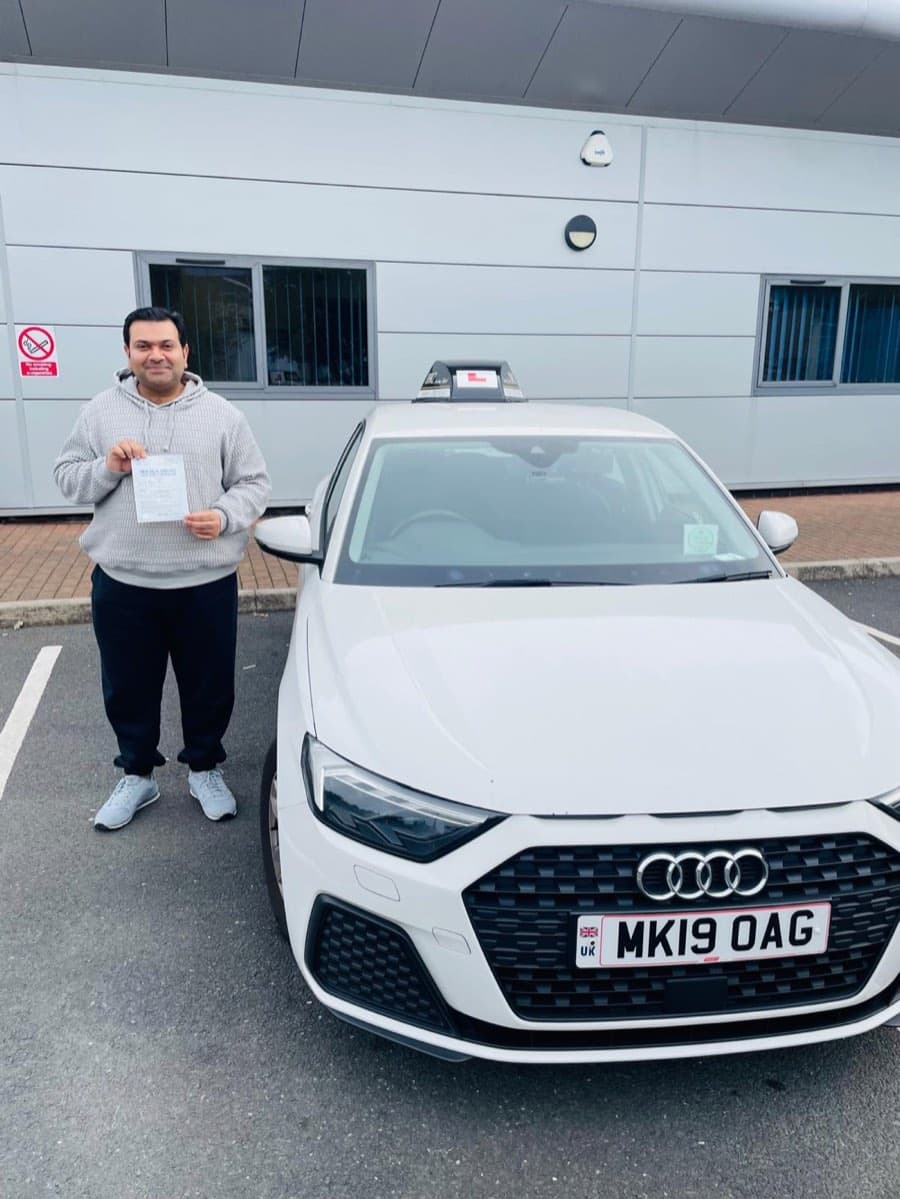 In a car park beside a modern grey building, a learner in a cream hoodie holds up a certificate by the bonnet.
