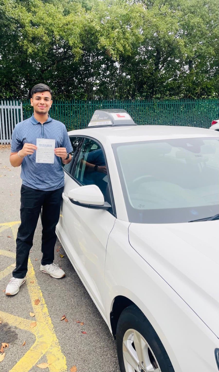 A learner in a blue polo shirt and dark trousers smiles with a certificate beside the tuition car, leaves on the tarmac.