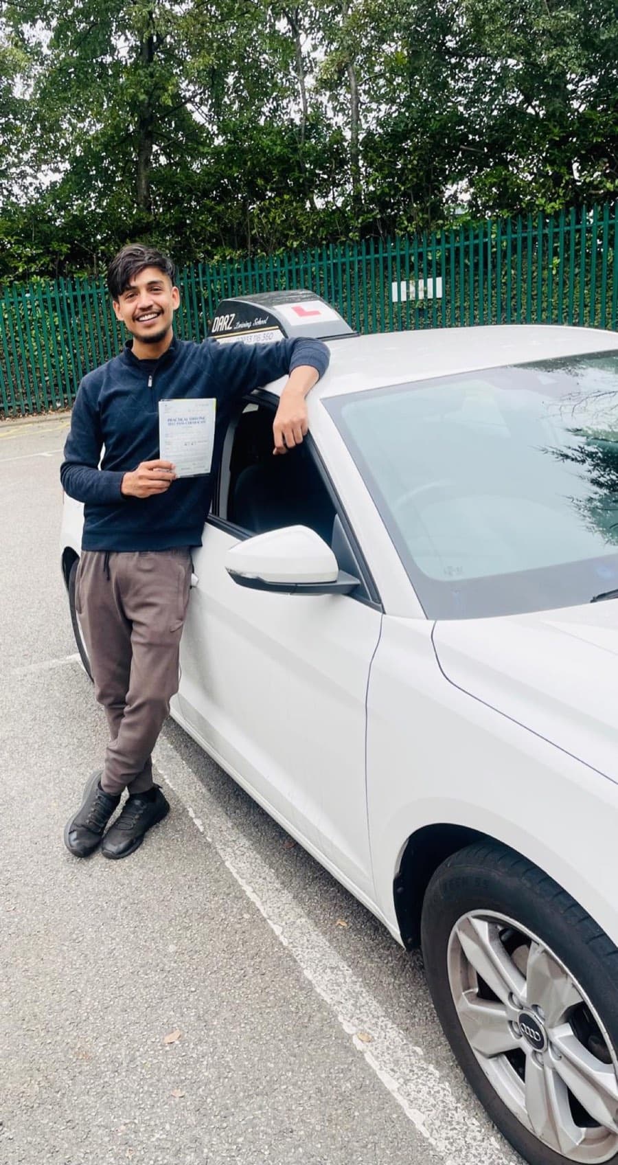 Resting an arm on the roof of the tuition car, a learner in a navy jumper displays a pass certificate.