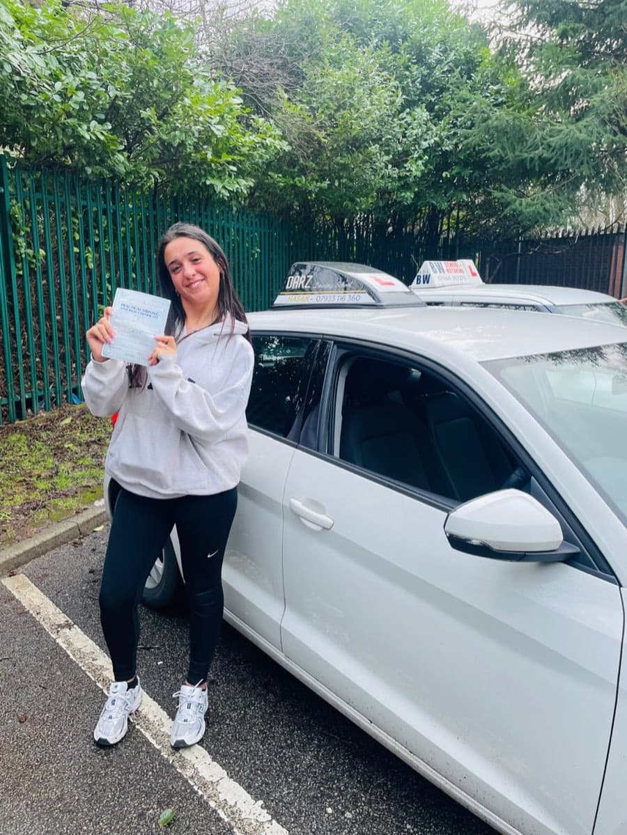 On a damp overcast day, a learner in a white hoodie raises a certificate beside the tuition car, another driving school car behind.