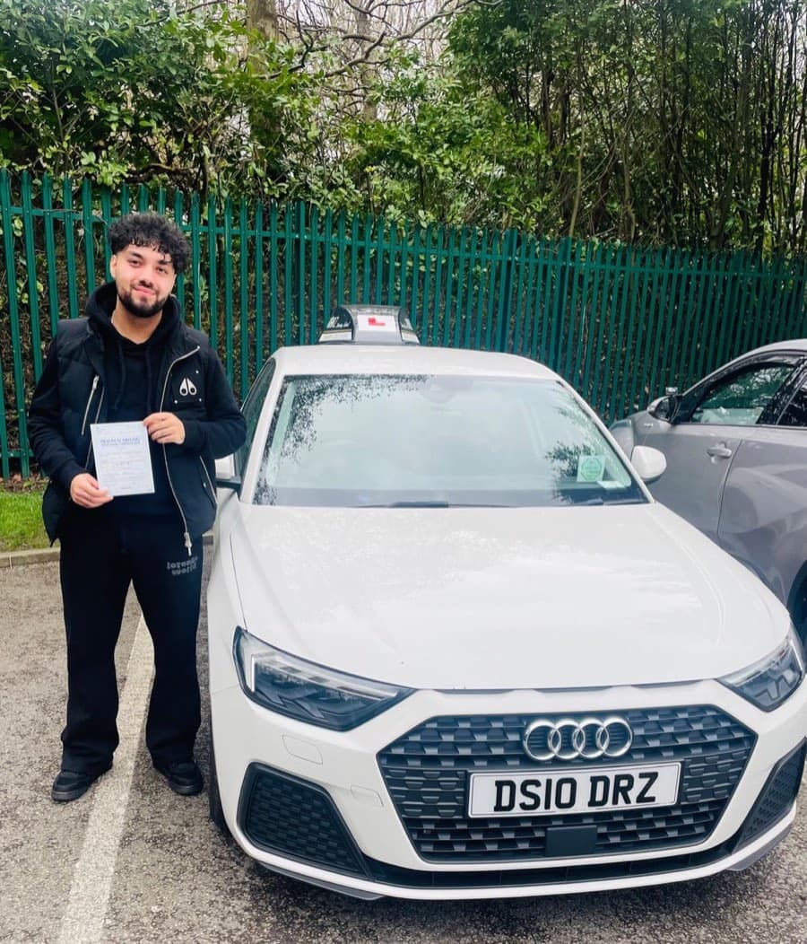 Standing at the front of the white tuition car, a learner in a black padded jacket holds a certificate.
