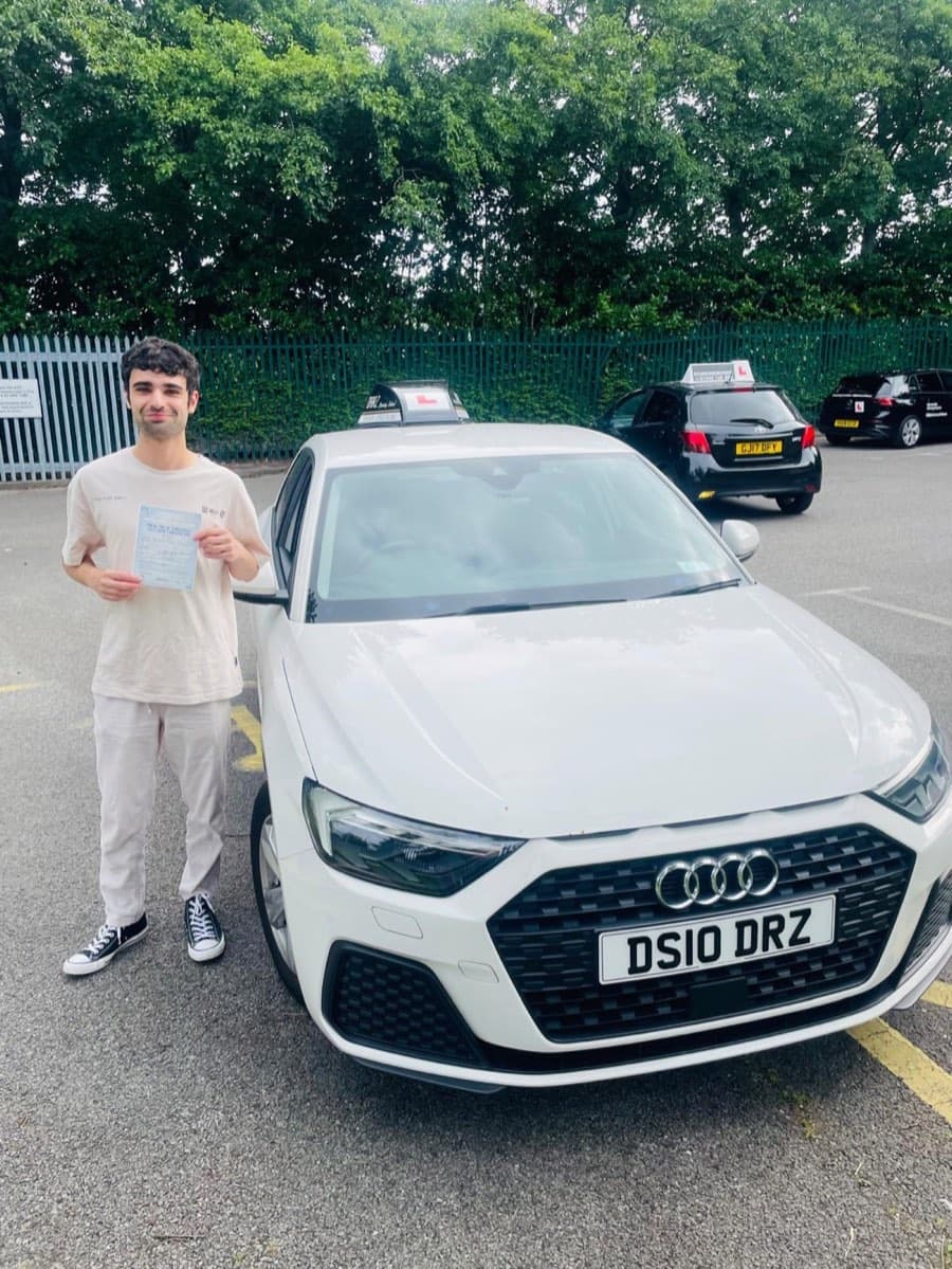 A learner in a cream t-shirt and pale trousers holding a pass certificate beside the white tuition car, green railings behind.