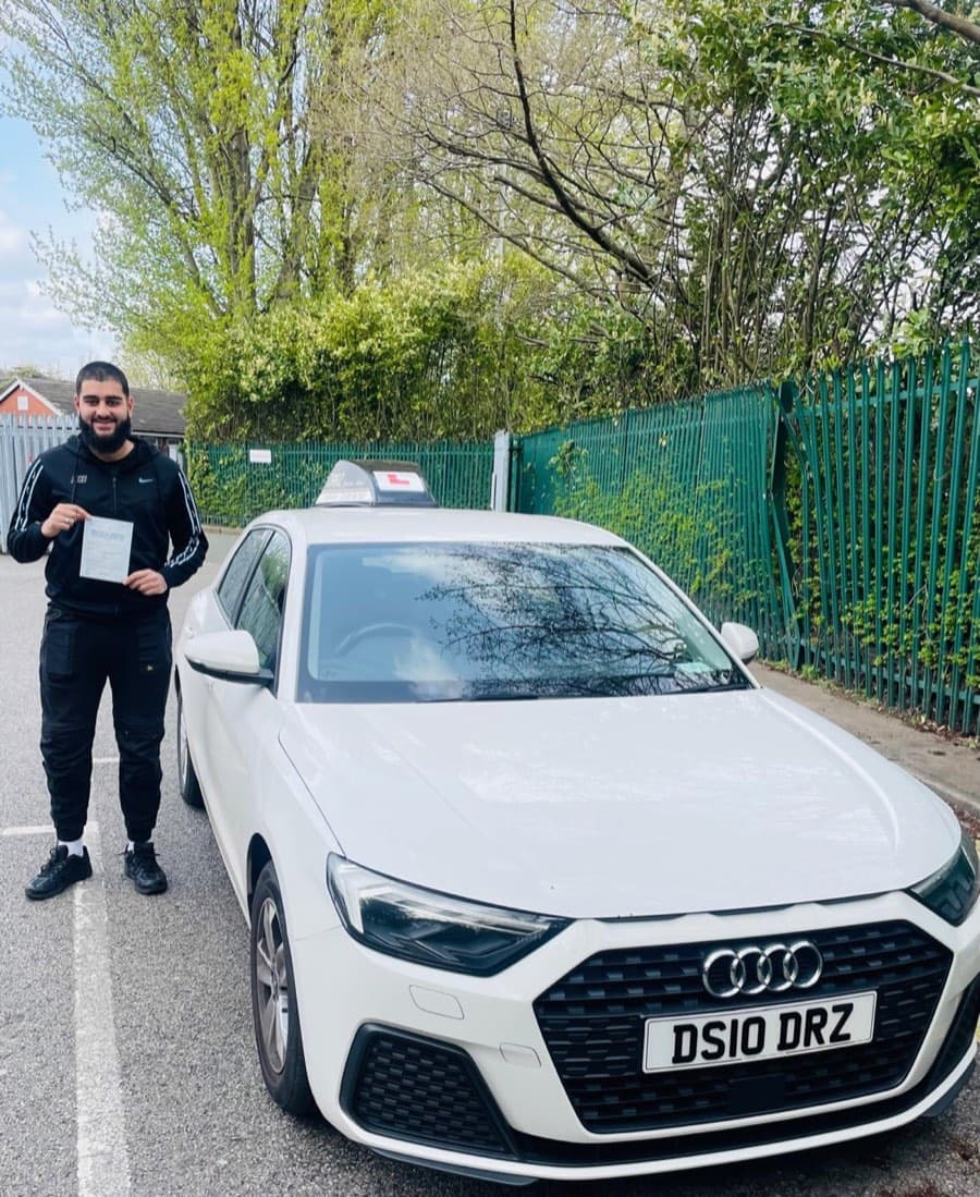A learner in a black tracksuit grins while holding up a certificate, green palisade fencing and spring foliage behind.