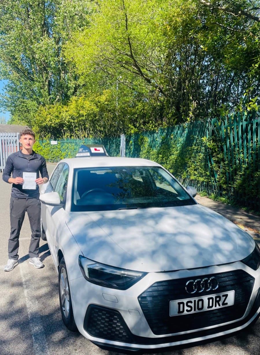 Dappled sunlight falls on a learner in a long-sleeved navy top and grey trousers presenting a certificate beside the tuition car.