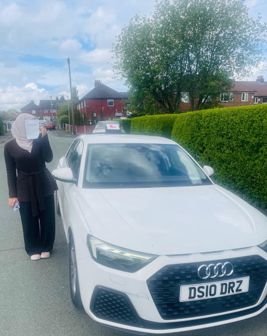 A learner in a headscarf and long dark cardigan holds the certificate up on a residential street of brick houses.