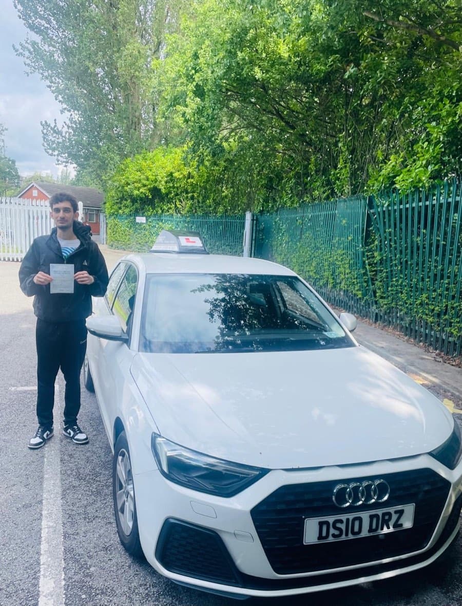 Certificate in hand, a learner in a dark jacket and black joggers stands by the tuition car beside a tall hedge.