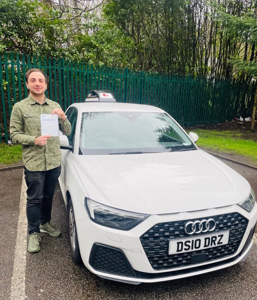 Wearing a green patterned shirt and dark jeans, a learner displays a certificate on damp tarmac by green railings.