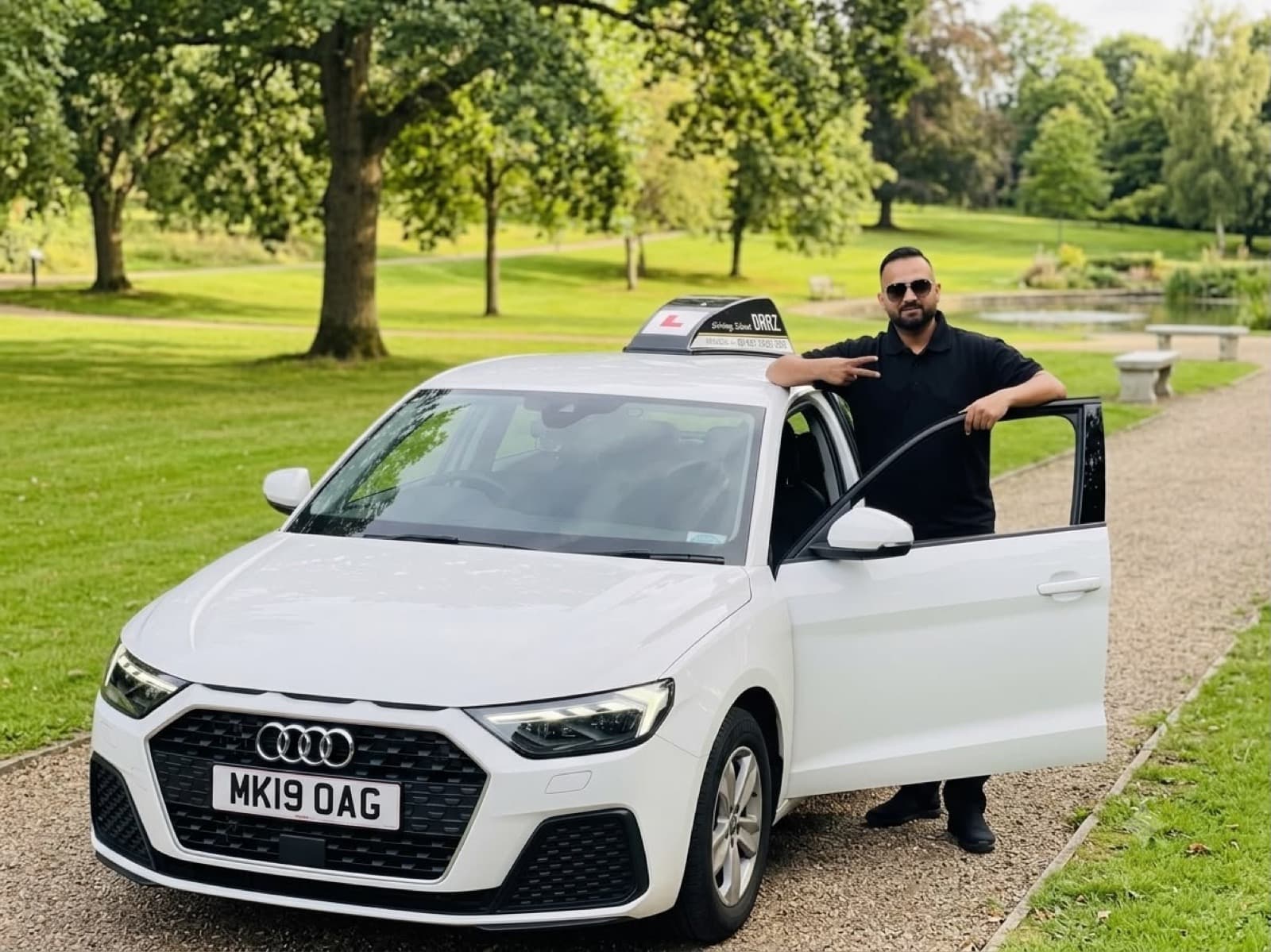 Hasan Ali standing beside the open driver's door of the white Magic Wheels tuition car, parked on a path in a Bolton park.